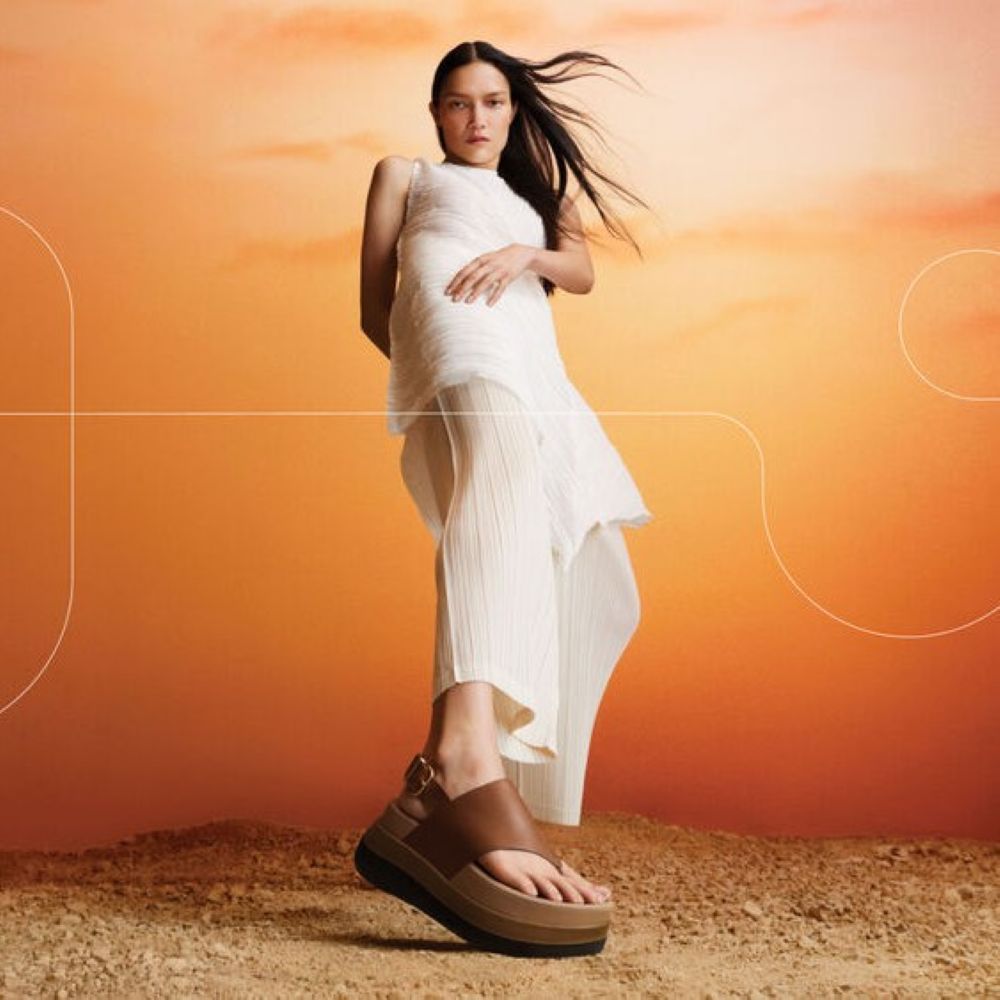 Model wearing brown wedge flip flop sandals sold at FitFlop in front of an orange background