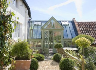 A greenhouse design painted in pale green bridging the gap between a white cottage and a terracotta tiled outbuilding, with gravel area and box hedges in front.