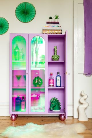 A pink display cabinet with iridescent glass doors decorated with vases, books and spirit bottles