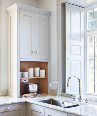 A built-in cabinet with paneled doors and round metal knobs sits above an open wooden shelf, which houses neatly arranged white ceramic canisters and cups. Below the shelf, a black toaster rests on the marble countertop beside a fresh loaf of bread.