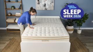 The image shows a dark haired mattress tester wearing a blue shirt and light trousers leaning onto a cream and gold mattress, set within a neutrally decorated bedroom. Overlaid in the top right hand corner a blue cloud-shaped badge reads 'Tom's Guide Sleep Week 2026'