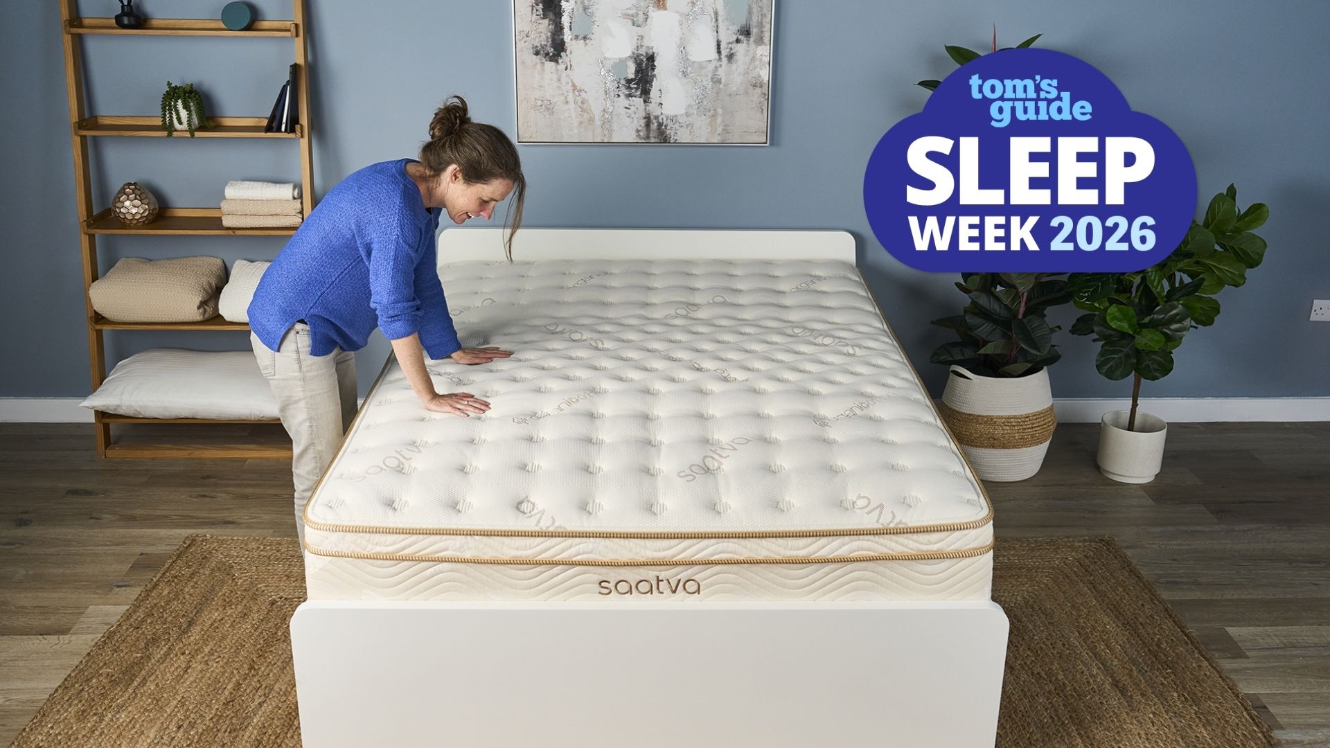 The image shows a dark haired mattress tester wearing a blue shirt and light trousers leaning onto a cream and gold mattress, set within a neutrally decorated bedroom. Overlaid in the top right hand corner a blue cloud-shaped badge reads 'Tom's Guide Sleep Week 2026'