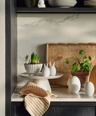 A white kitchen counter with Easter decorations on a cake tray, beside some freshly sprouting plants and flowers.