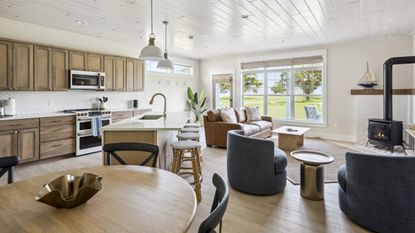 Large open-plan space with kitchen island and stools; kitchen area with oven/stove cooker, wooden cabinets and microwave; living room area with sofa, armchair and small coffee table; large, round, wooden table in the foreground. All on wooden floor with bright windows in background.