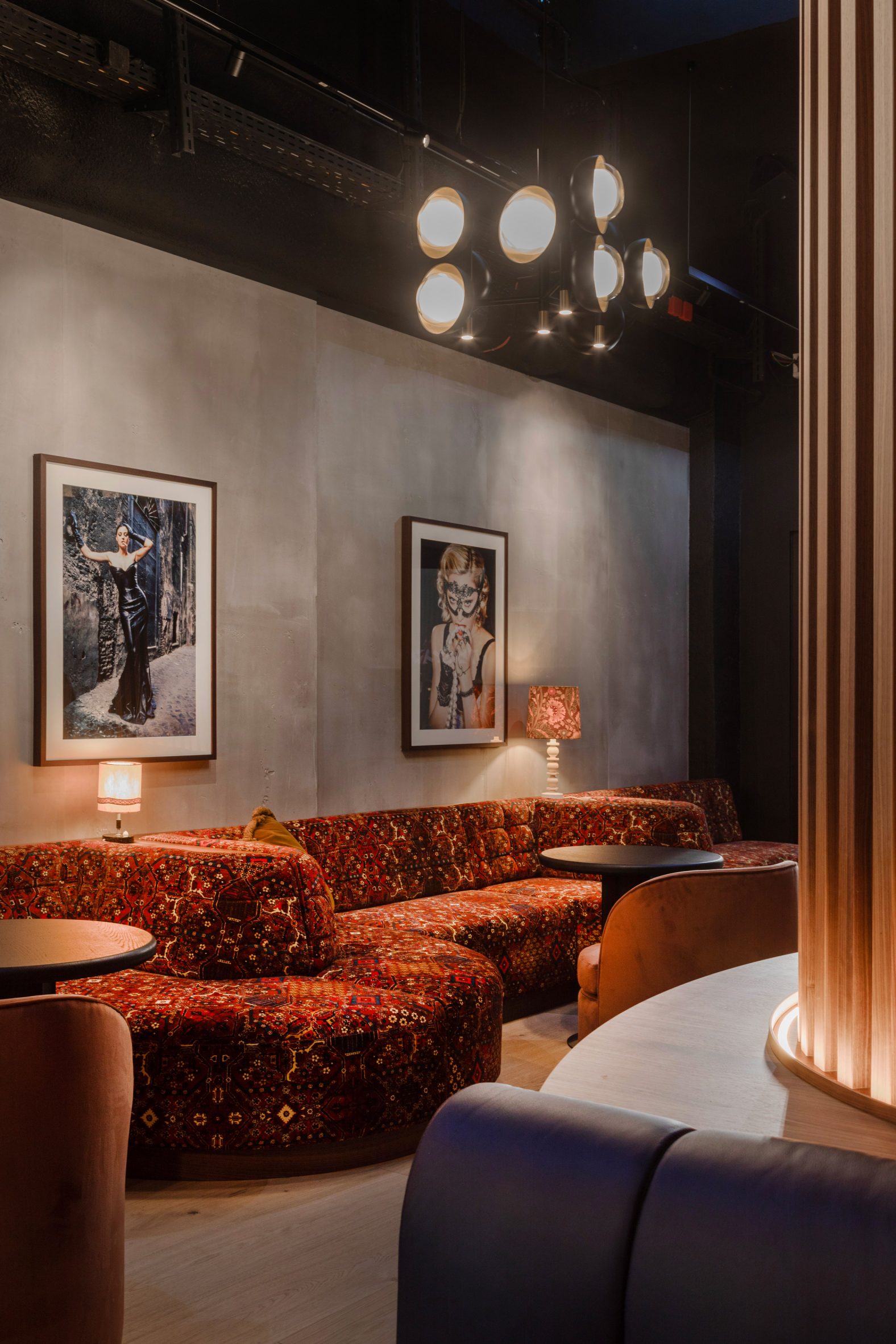 A dimly lit bar with texturally upholstered red banquettes, warm table lamps, framed photographs hanging on cement walls, and round wooden tables.