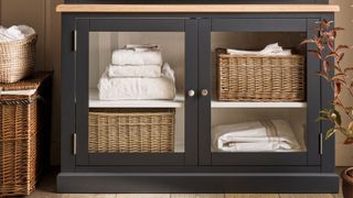A dark charcoal grey glazed linen cabinet with an oak top, shelves holding folded white towels and rattan baskets, with wicker laundry baskets beside it and a burgundy plant alongside.