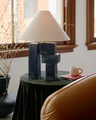 A minimalism-inspired blue and white table lamp sits on top of a round table covered in green velvet next to an espresso coffee cup in white.