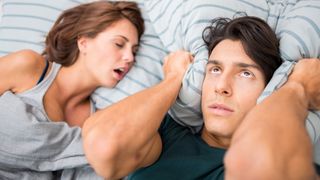 A man with dark hair rolls his eyes in frustration and holds a pillow to his ears because his partner is snoring