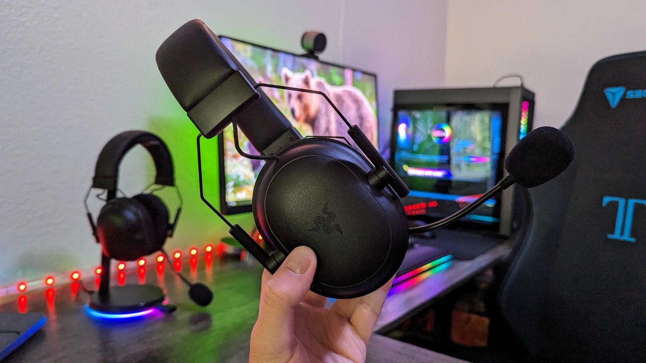 A hand holds a black gaming headset with a microphone in a setup featuring colorful LED lights. A monitor and a gaming chair are in the background.