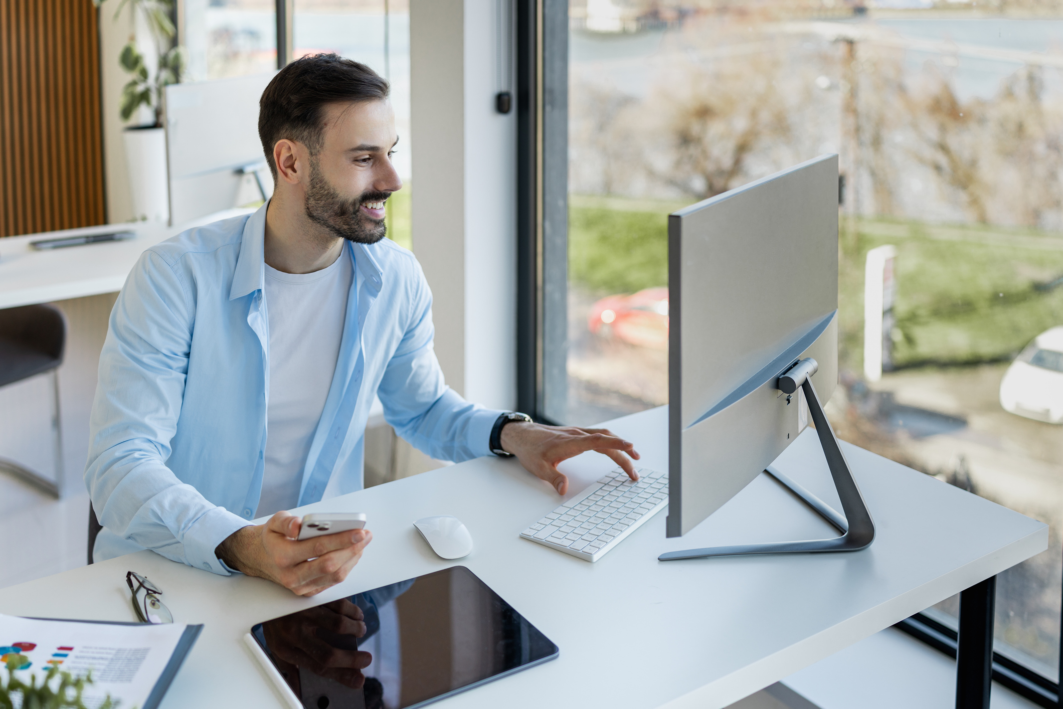A man in a light blue shirt smiles while working at a computer in a modern, bright office with large windows overlooking a scenic outdoor view.
