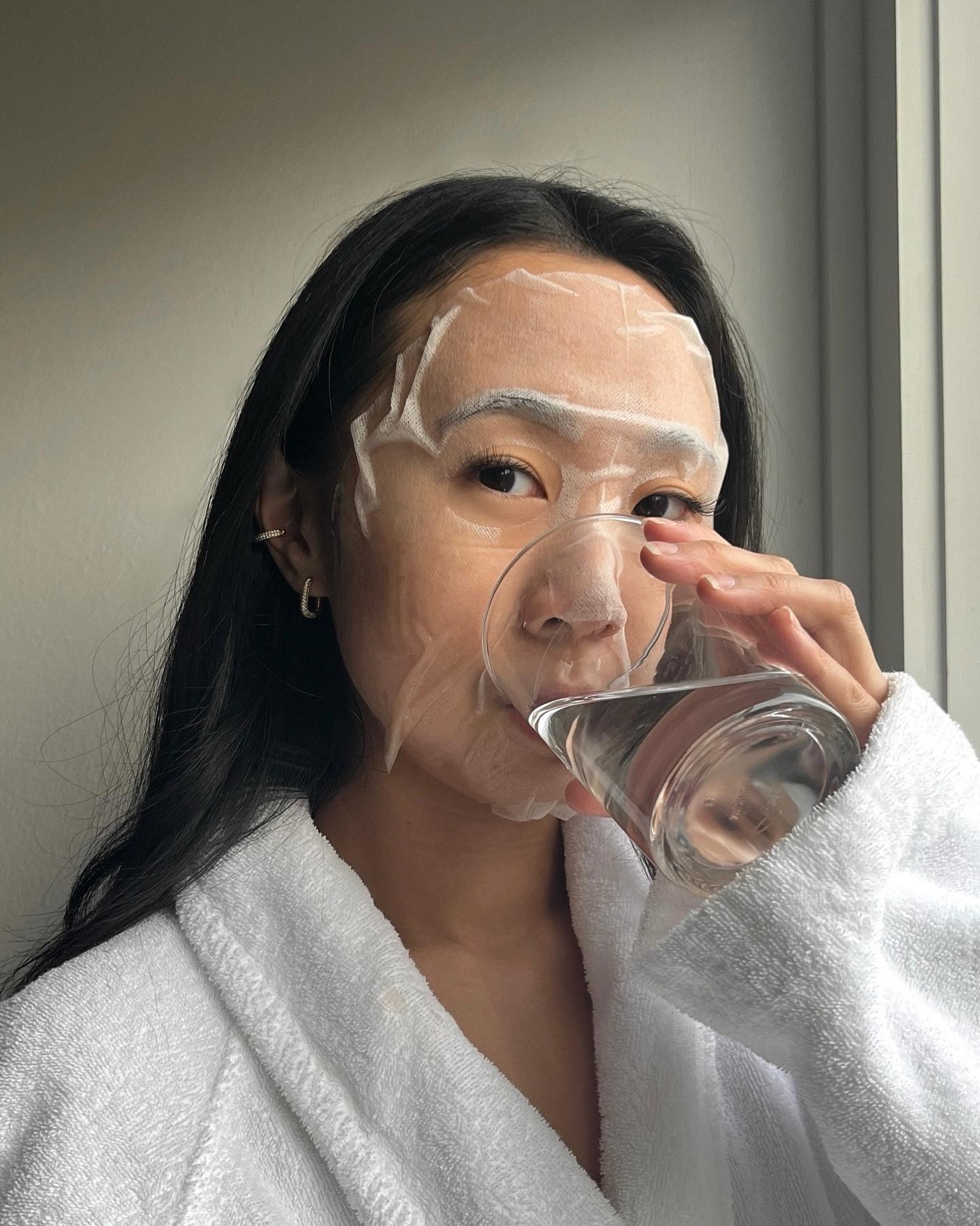 A woman wearing a sheet mask drinking a glass of water