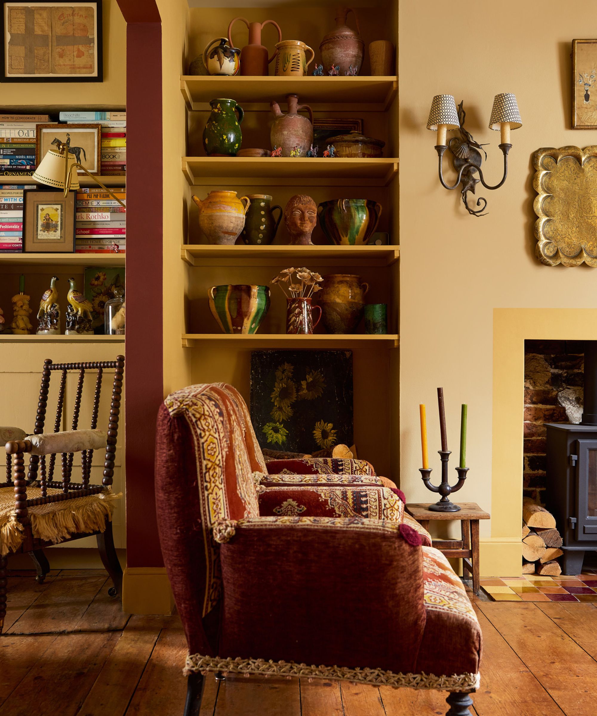 Mustard yellow living room with shelves of antique ceramic pots, a wood-burner, a pair of vintage burgundy tapestry armchairs, wooden floors, and a doorway painted burgundy.