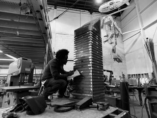 Black and white image of Arrechea in his white brick wall workshop, knelt at his 20ft stainless steel construction, surrounded by equipment, tools, lighting and work benches