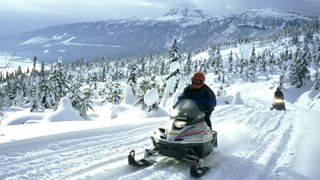 two snowmobilers on a very snowy pathway with pine trees in behind. far in the background are mountaintops and cloudy sky