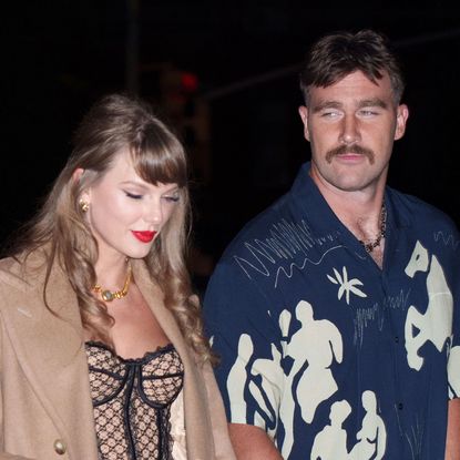 Taylor Swift wearing a tan coat and bustier walking at night next to Travis Kelce who is wearing a blue wildly patterned shirt