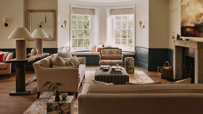 neutral living room with original stone fireplace, two tone half panel in navy blue, neutral couches, a couch table with two large lamps and a striped ottoman