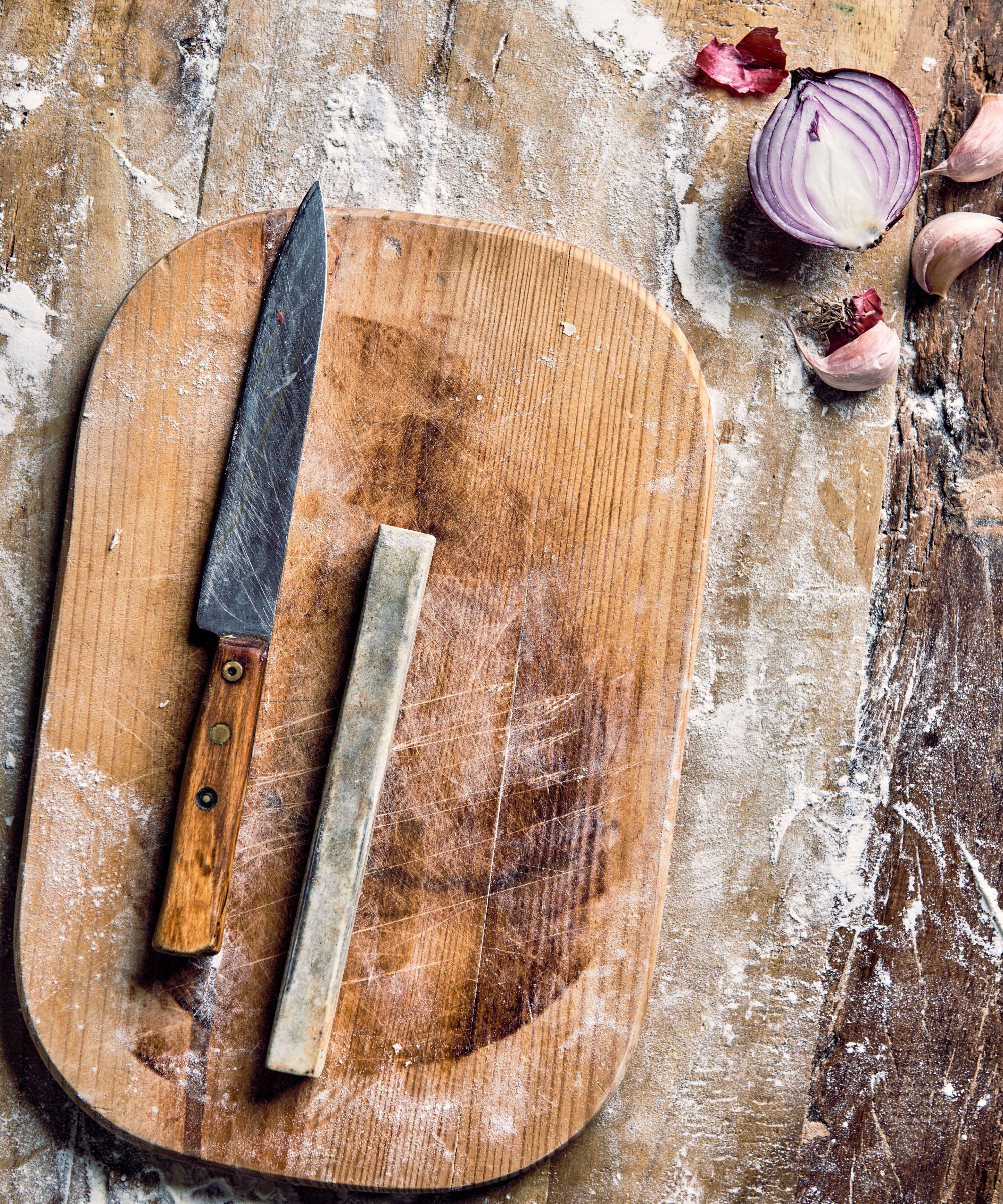 A wooden handled knife and thin whetstone on an oval chopping board, on top of a wooden surface covered in flour, with half a red onion and three garlic cloves seen in the top right corner.