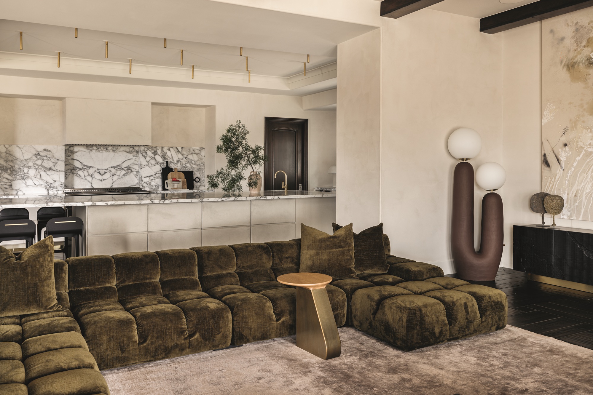 modern living room with velvet dark green sofa, light brown rug, sculptural floor lamp, beige kitchen in background with marble backsplash and dark timber bar stools