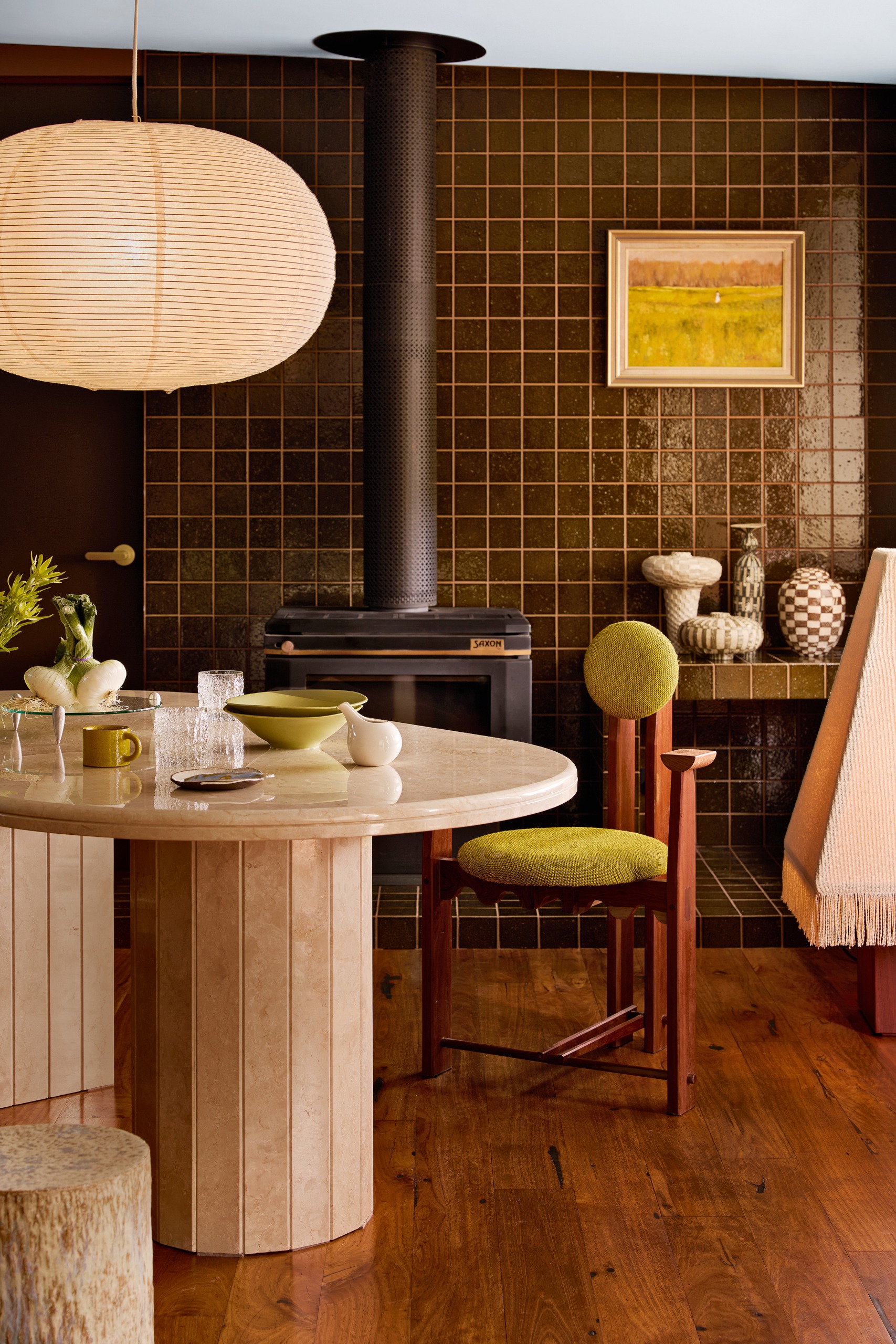 Dining room with round travertine table, red wood and green upholstered chair, black woodburner on a platform of dark olive green zellige tiles, and a paper pendant light