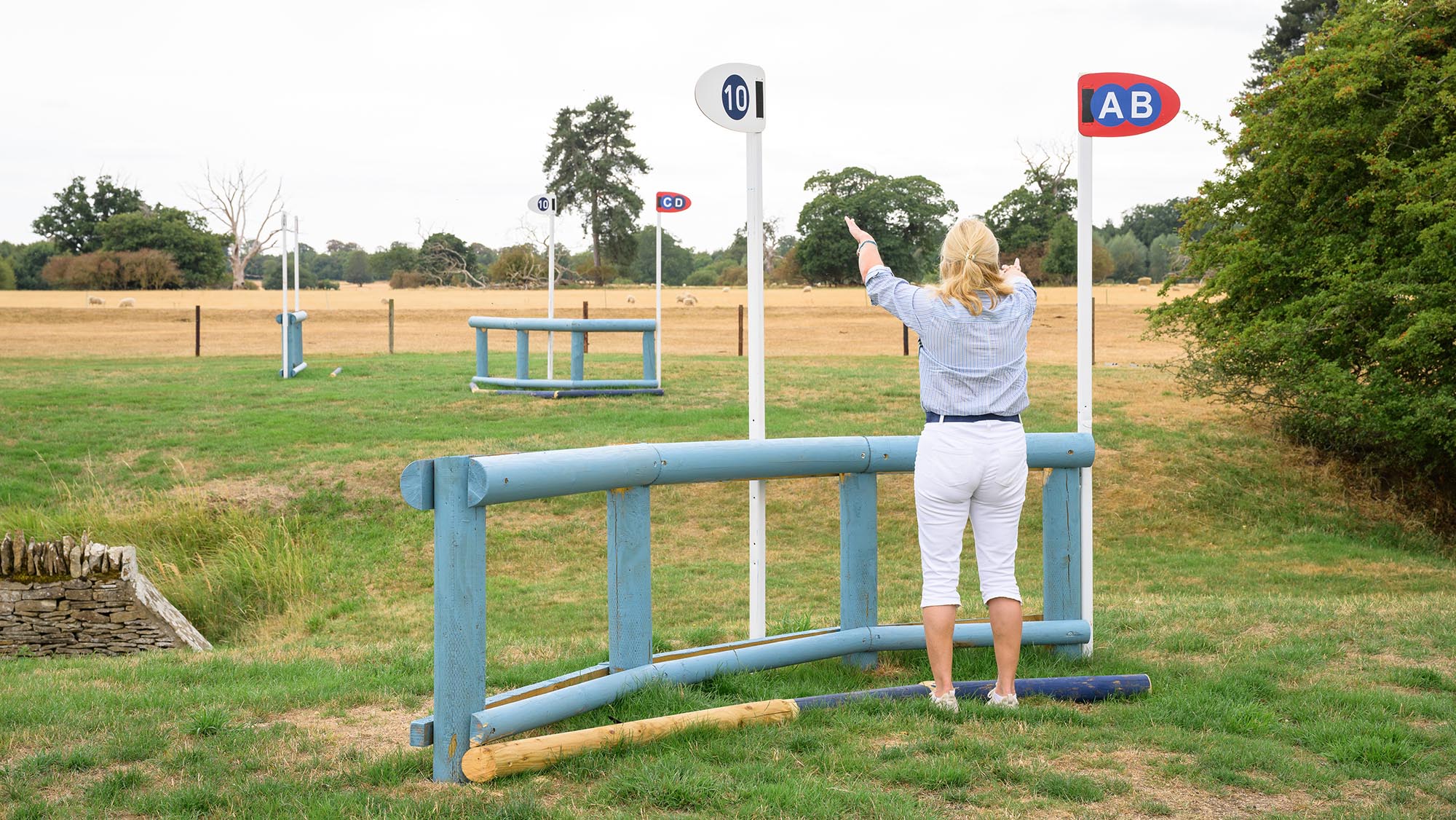 Back view of event rider Tina Cook with her arms up, standing in front of a pale blue fence frame, with another fence in the background, on the Burghley cross-country course 2026