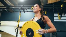 A smiling woman stands in a gym holding a yellow weight plate. Behind her we see a metal pull-up bar draped with TRX straps.