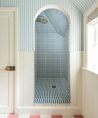 Walk in shower with blue stripe tiles, arch, pink checked floor tiles, beige stripe blind, shiplap painted white