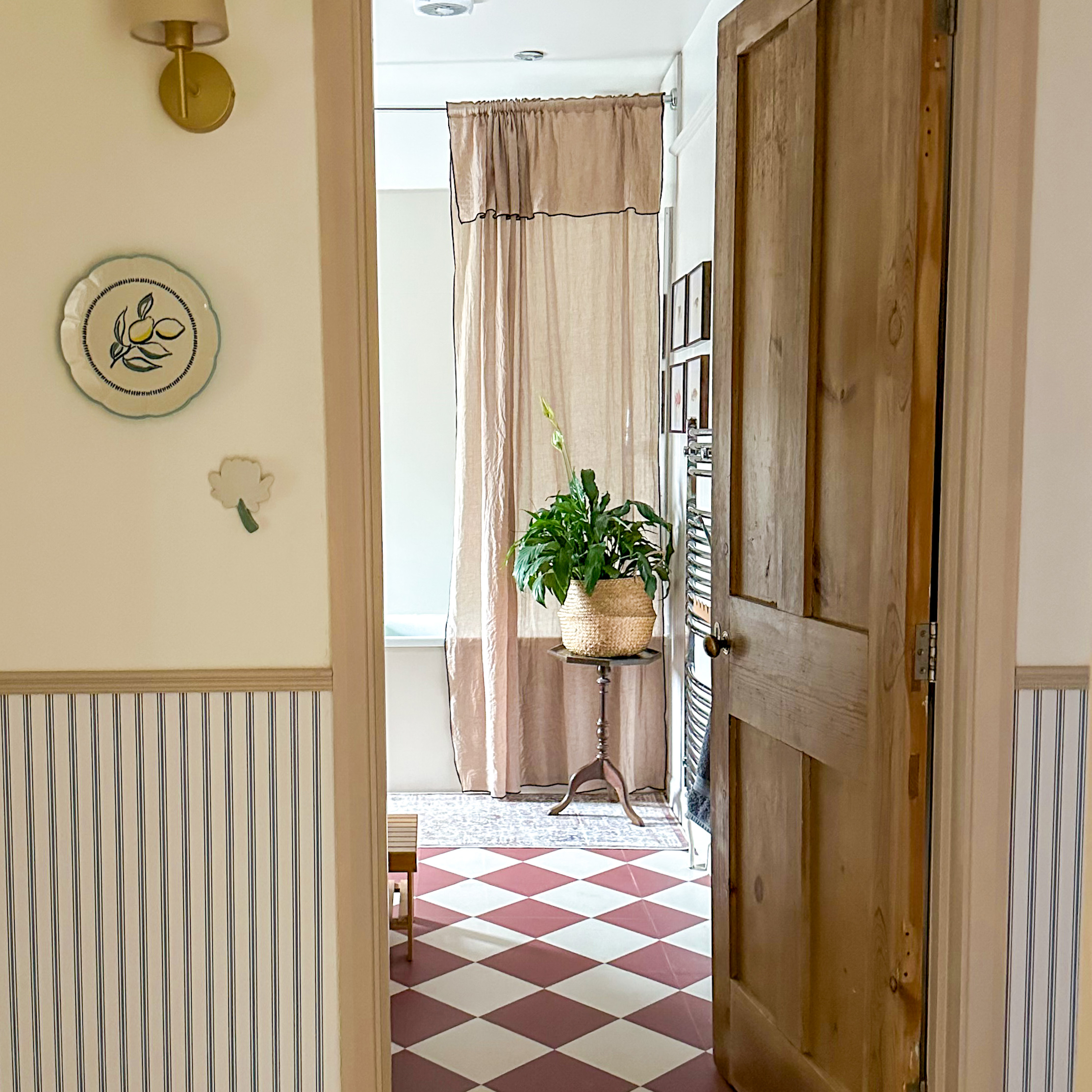 looking through a wooden internal door from a neutral hallway into a bathroom with red tiled floor and beige shower curtain