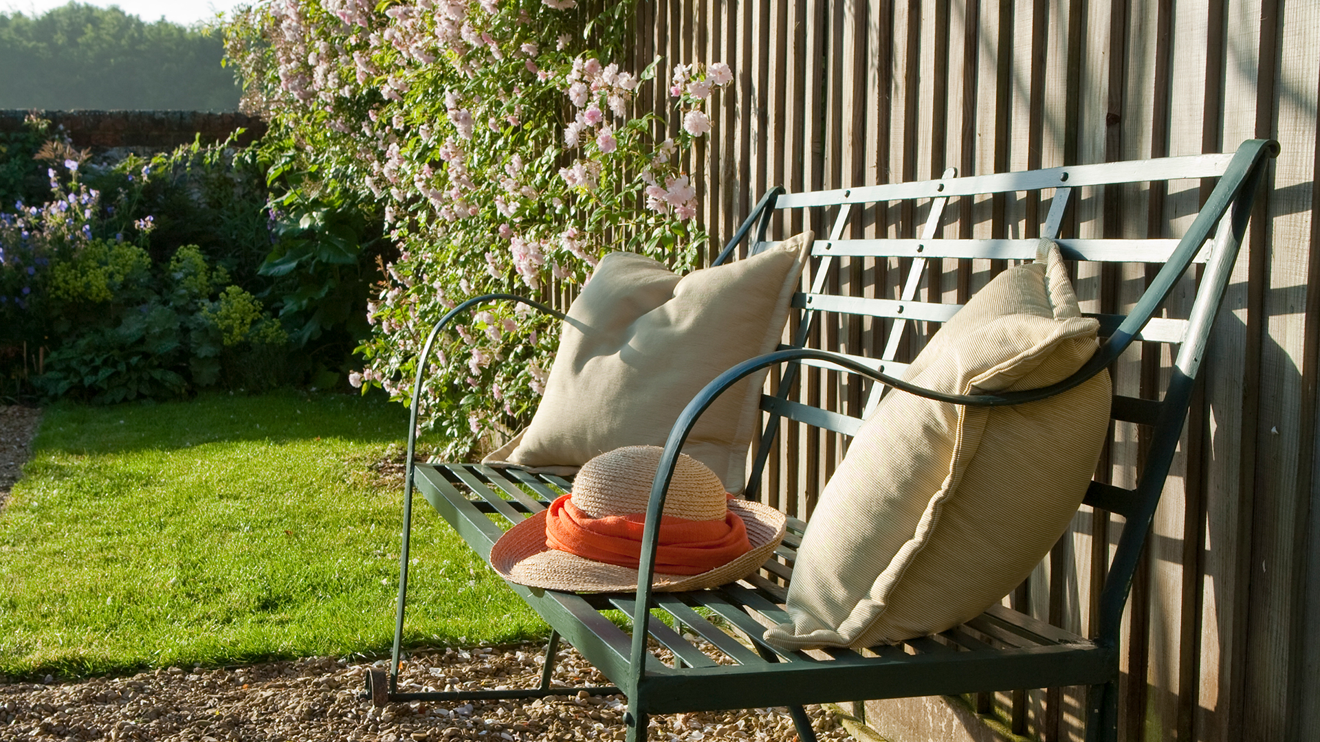 garden with a wire bench with cushions and a sun hat, pushed up against a wooden garden fence
