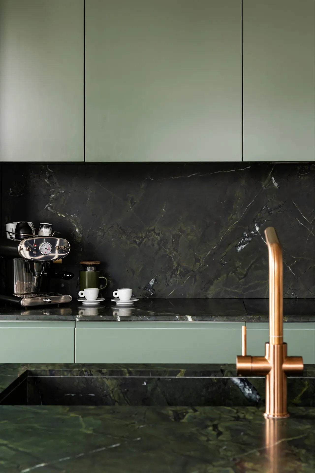 Image of a kitchen with light green cabinetry and a dark green marble countertop. There is an espresso machine on the counter, along with two white espresso cups.