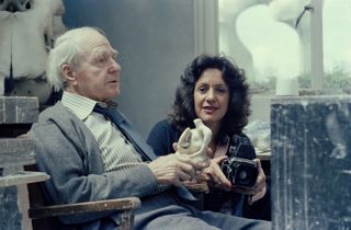 An old man dressed in a stripy shirt, tie, and grey cardigan sits in a wooden chair in a studio while holding a clay statuette next to a woman wearing a blue cardigan, a stripy red and white shit, and holding a medium format camera.