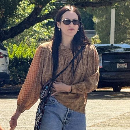 Riley Keough walks down the street wearing straight-leg jeans with a blouse and high-vamp flats.