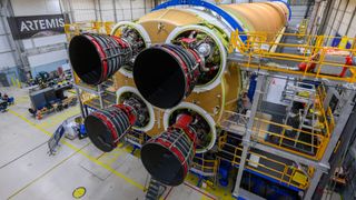 a golden rocket stage and its four black engines are seen in a horizontal position inside a large warehouse-like facility.