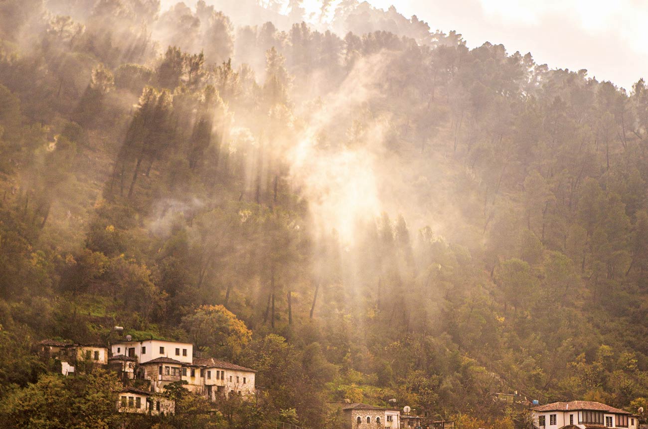 Shafts of sunshine emerge from the mist over a steep hill, with houses on the hillside
