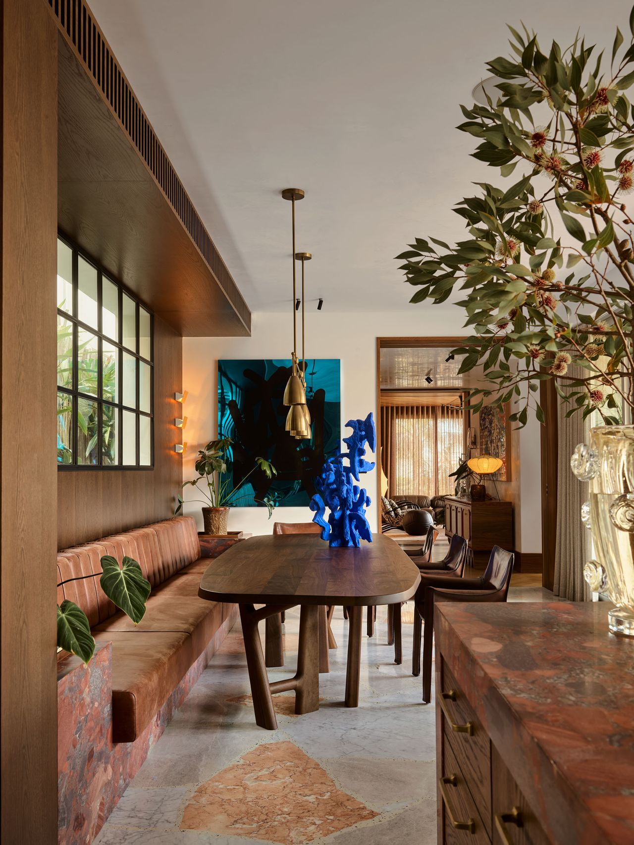 dining banquette area with marble floors, timber table, artwork, hanging pendants and corner of kitchen island visible with vase filled with large branches