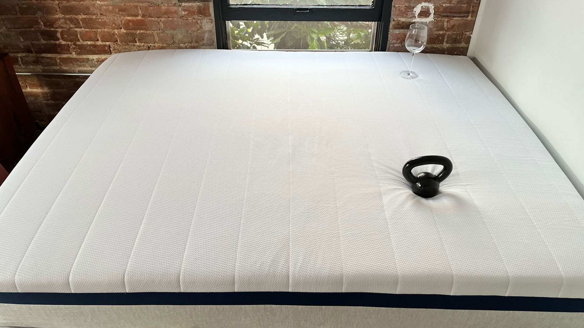 Photo of Helix Midnight mattress in a bedroom, with a weight on it, and a wine glass stood upright on the mattress surface nearby