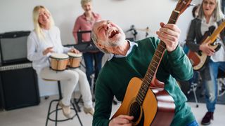 A group of senior musicians is led by a man in a green jumper, who is leaning back with his moth open as he plays a dreadnought acoustic