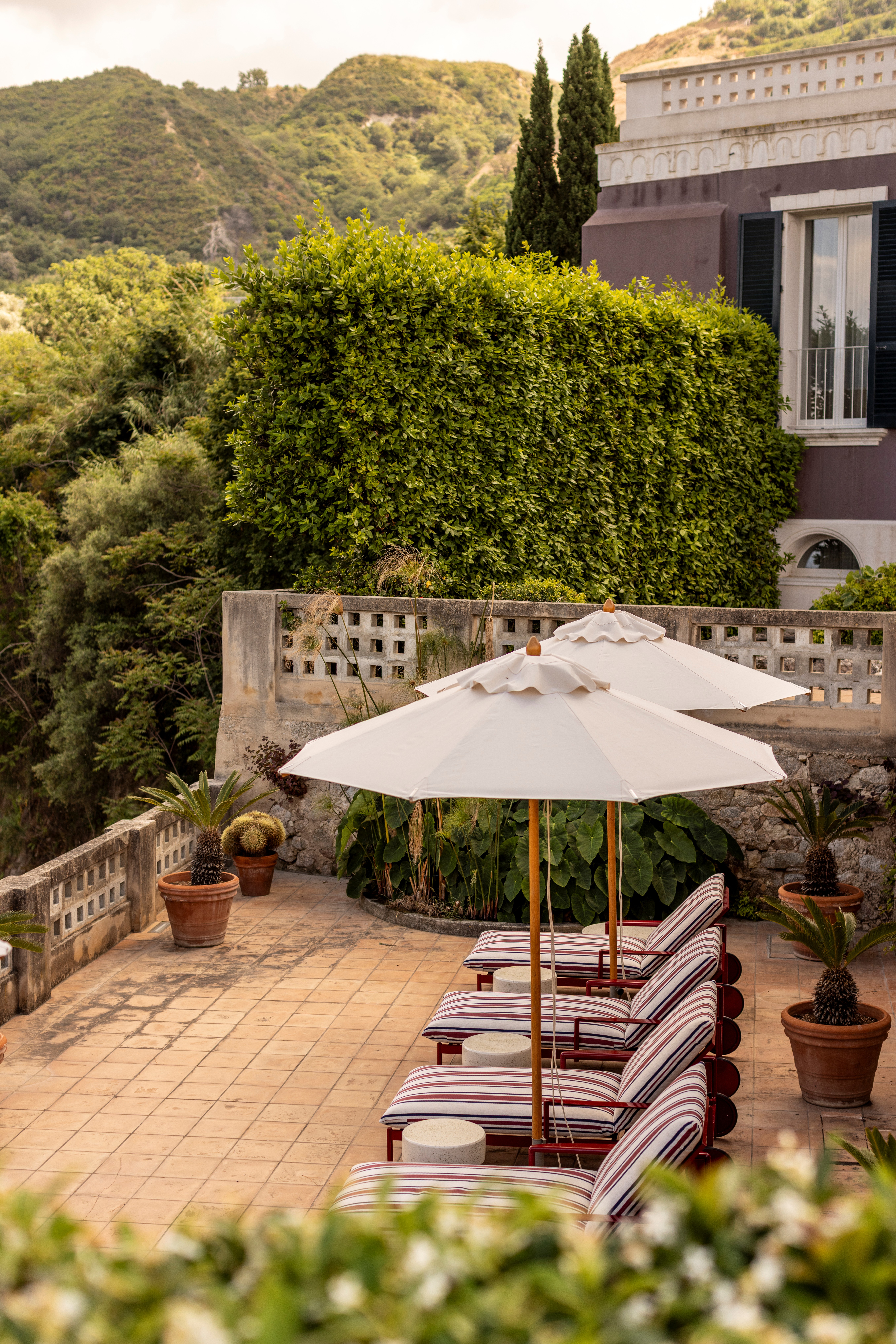 A Mediterranean hotel wrapped in raw greenery and with historical architecture houses nudely painted rooms with linear, warm decor, while, outside, stripes and vintage parasols set a nostalgic vibe.