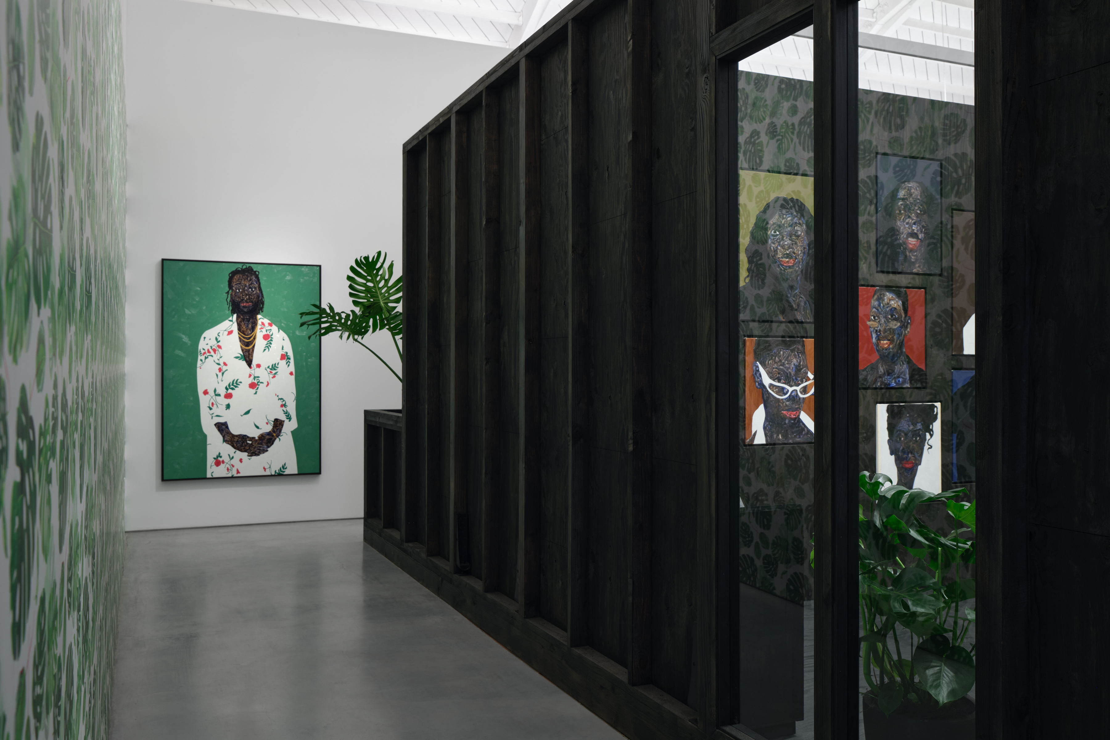 View of an art gallery installation with dark wood walls and multiple expressive portrait paintings. In the foreground, a large black metal structure with vertical bars frames additional paintings inside, while more portraits hang on the walls in the background.