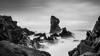 A dramatic black and white coastal scene featuring jagged rocks and a towering rock formation, with gentle waves lapping at the shoreline