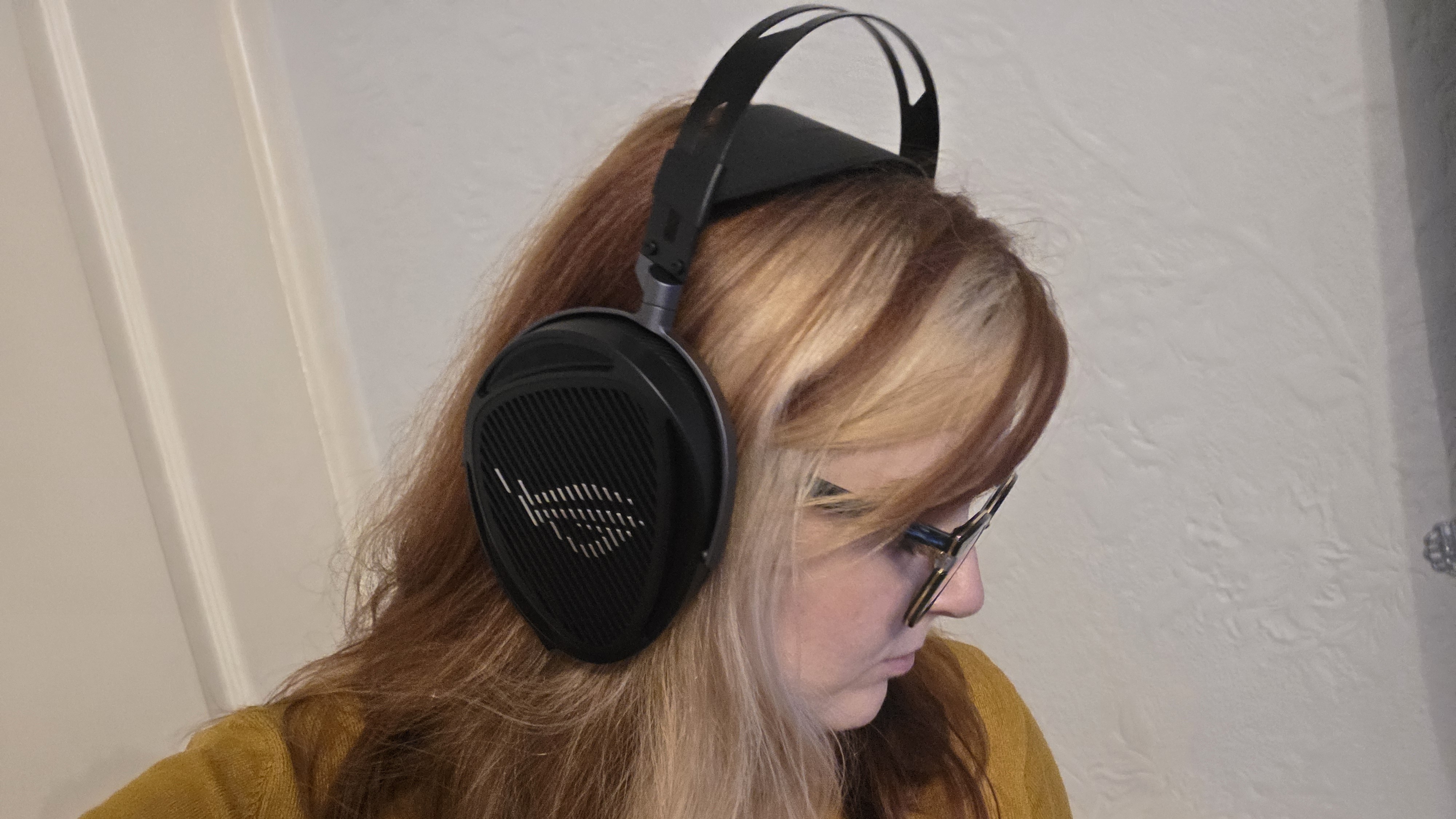 A person with long, blonde and auburn hair wears large over-ear headphones. They are looking down, wearing glasses and a mustard yellow sweater.