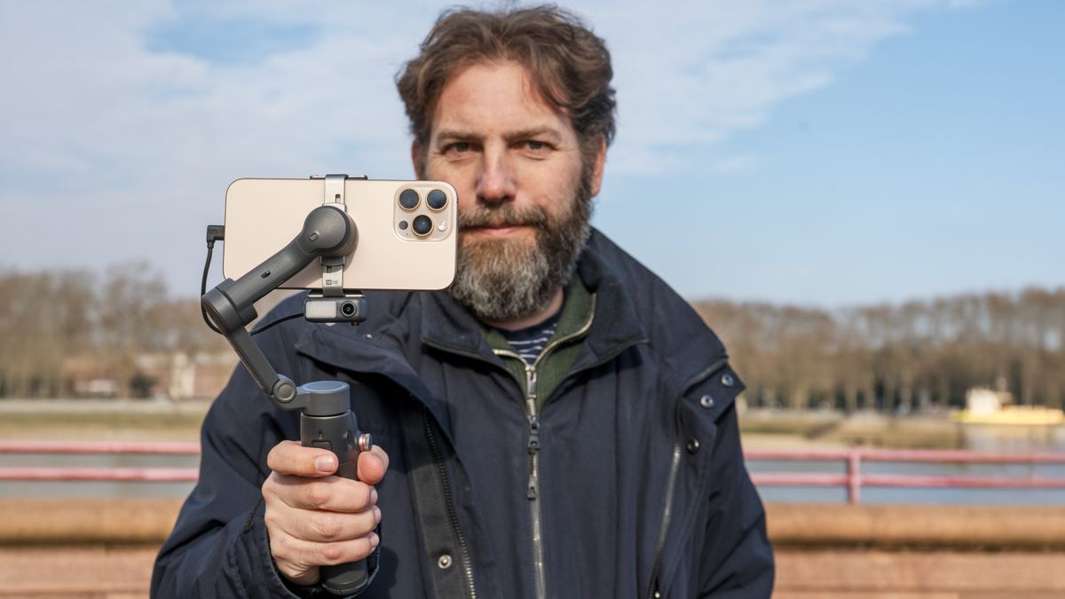 Osmo Mobile 7P in Adam Juniper&#039;s hand by river Thames with blue sky behind and iPhone 16 Pro Max in grip