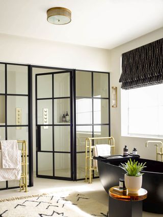 black and white bathroom with black cristal shower doors, black tub, gold towel rails, black and white rug and blind