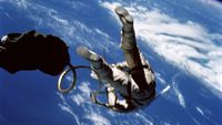 an astronaut in a white spacesuit is seen from below and behind while on a spacewalk outside his space capsule. Earth below serves as a backdrop.