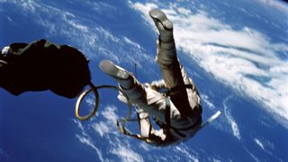 an astronaut in a white spacesuit is seen from below and behind while on a spacewalk outside his space capsule. Earth below serves as a backdrop.