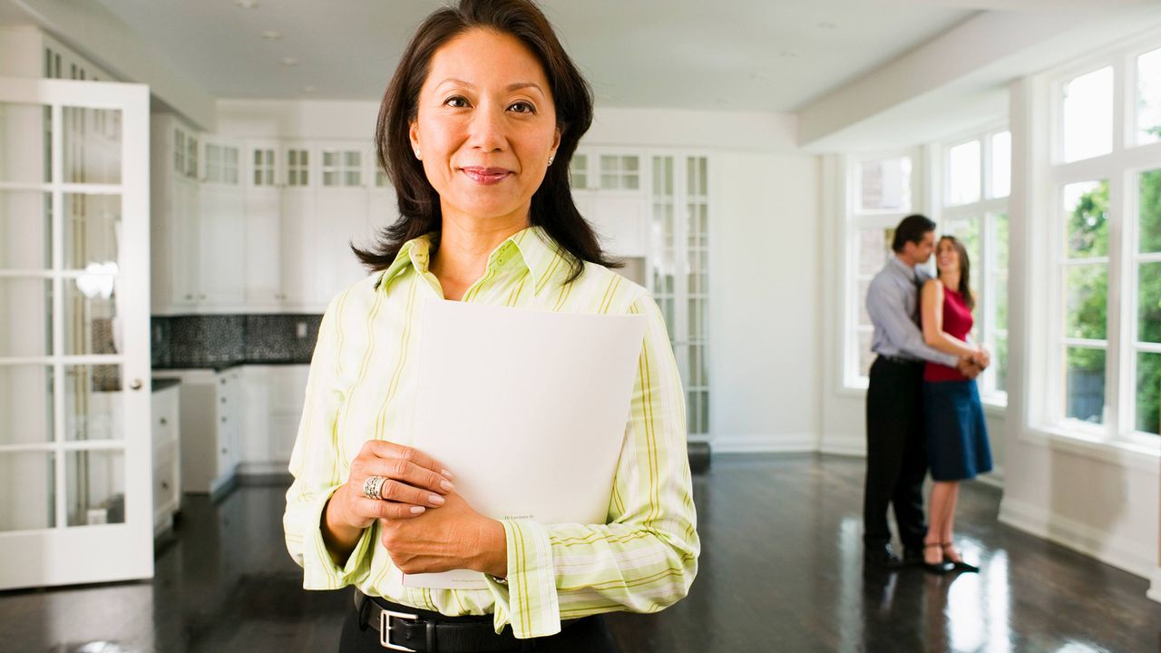 A Realtor stands in an empty living room with a happy couple in the background.