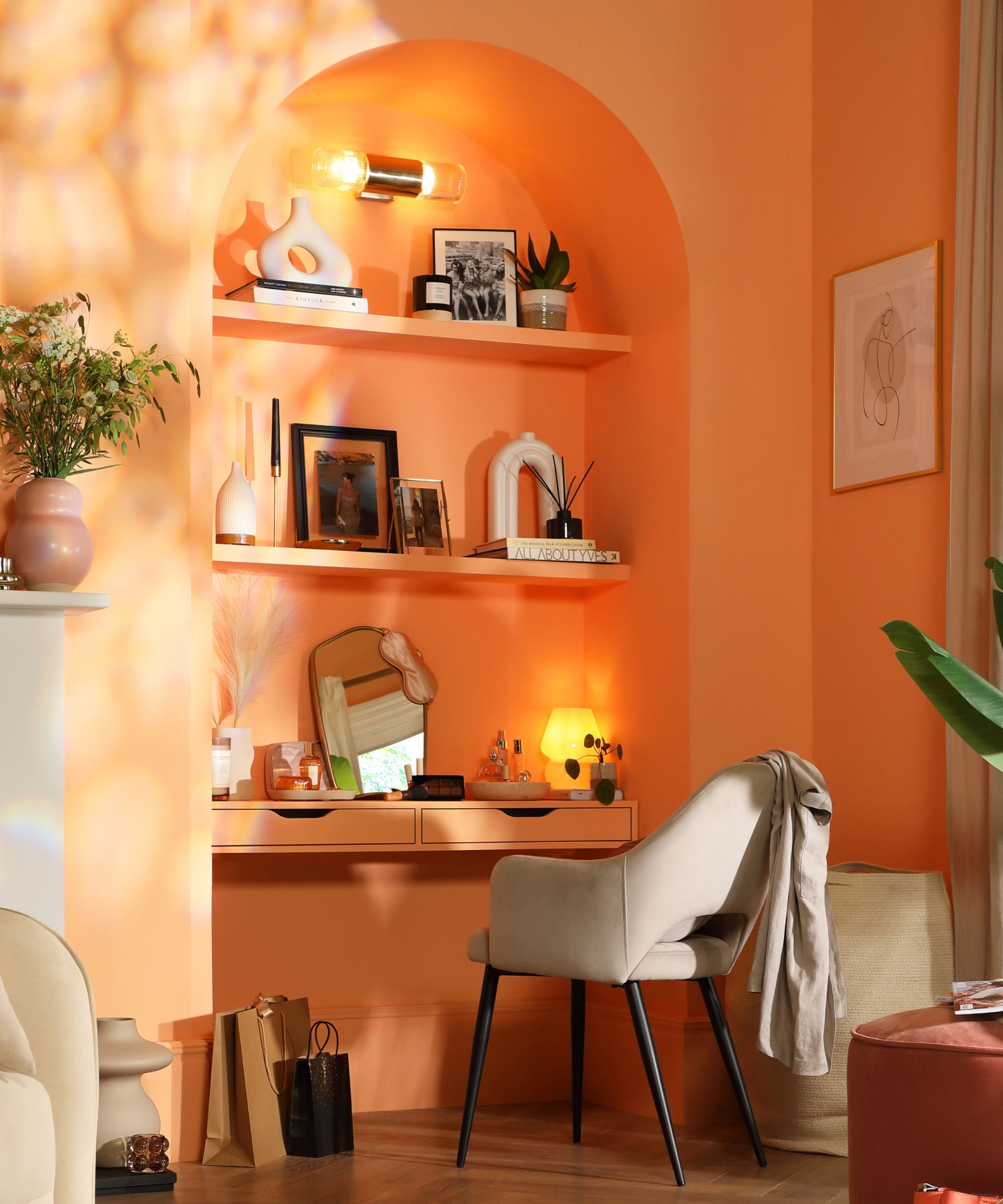 A cosy alcove painted in a warm peachy-orange tone features built-in shelves styled with books, framed photos, vases, and decorative objects. A small desk with drawers is set into the alcove, topped with a mirror and table lamp.
