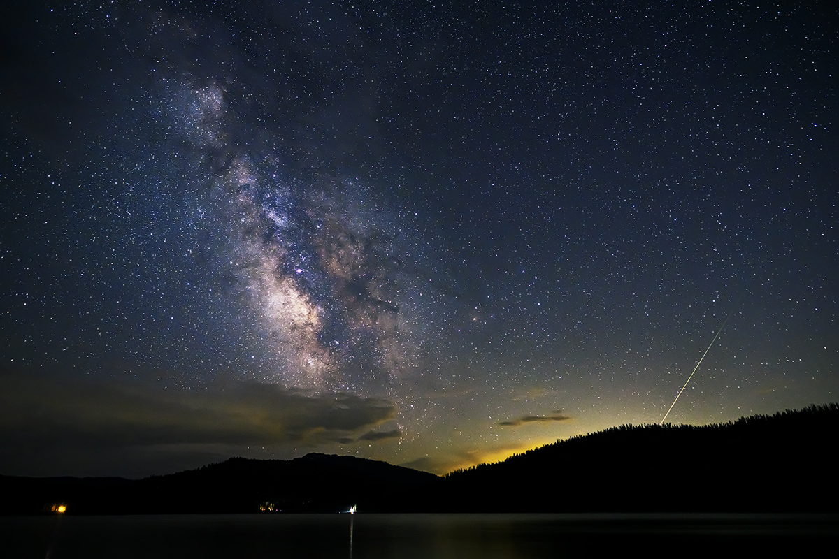 Uma chuva de meteoros é retratada contra um céu estrelado e um primeiro plano cênico