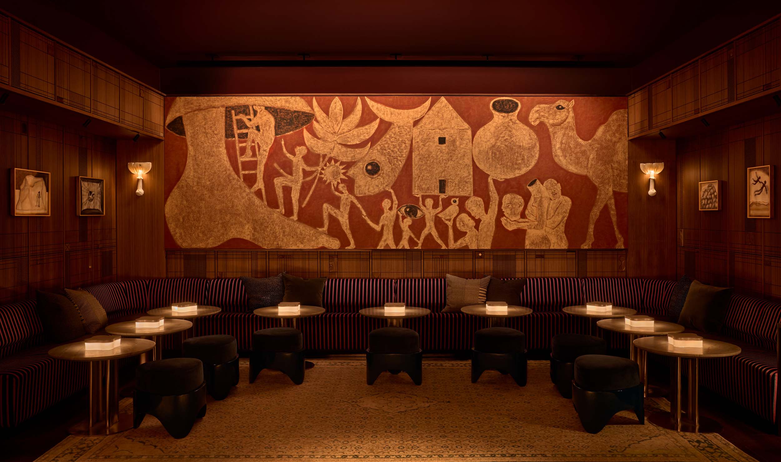 A beautiful, archaic art-inspired mural in the tones of terracotta red, pale pink, and black depicts stylized humans queing to climb up a ladder positioned at the base of a large foot in a warmly lit bar filled with round coffee tables and soft banquettes.