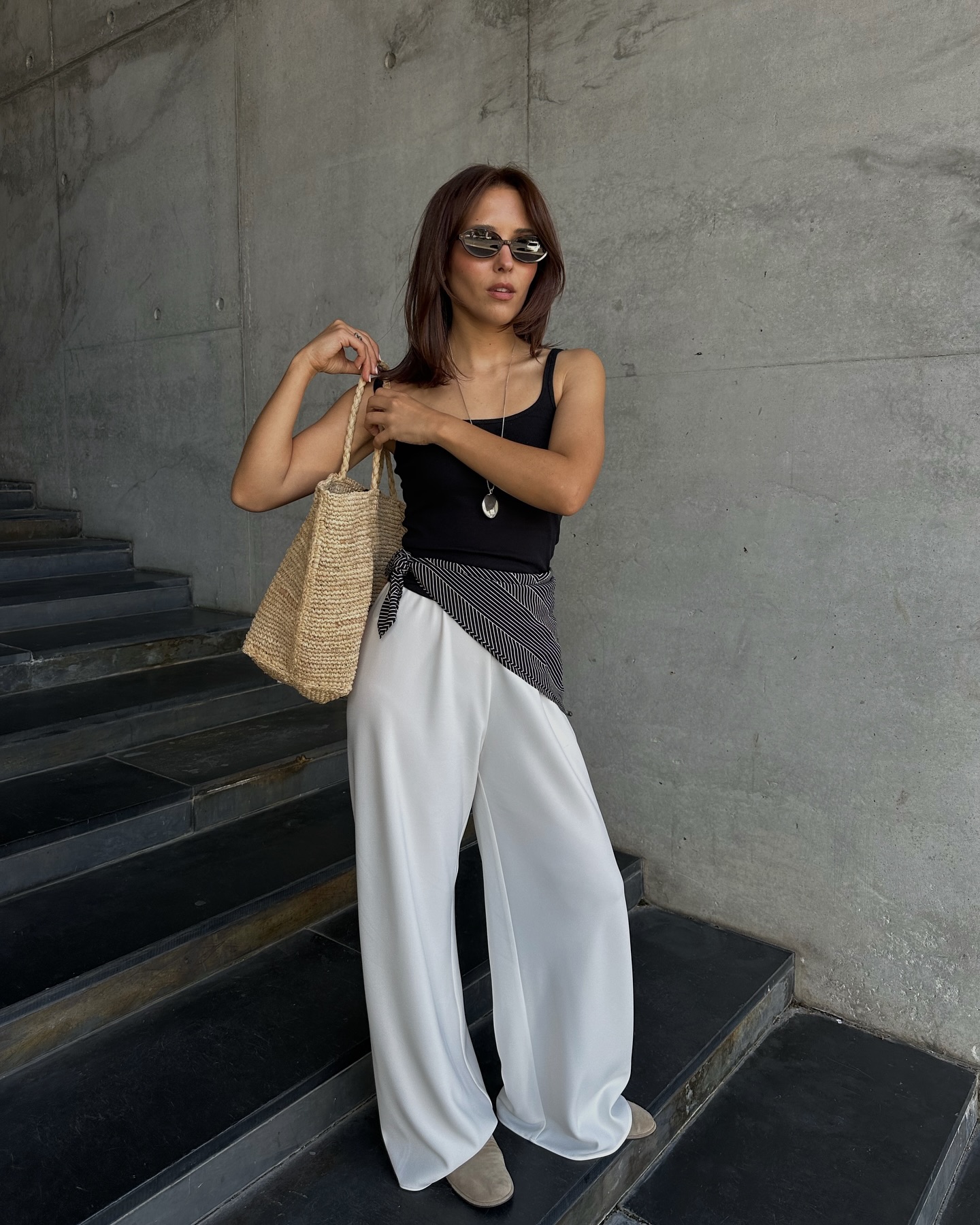 An image of @deborabrosa wearing a holiday outfit consisting of a black tank top, white silk pants, scarf belt and raffia bag.