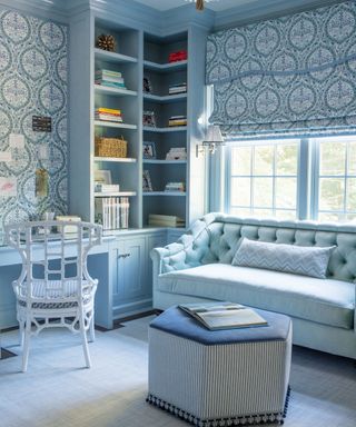Home office in blue and white with patterned wallpaper and blind, painted desk and shelving, sofa and ottoman